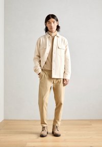 Beige shirt jacket with two front pockets, beige turtleneck, and light beige pants, complemented by brown lace-up boots.