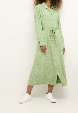 Frau trägt ein langärmliges, grün gemustertes Midikleid mit gebundenem Taillengürtel und weißen Turnschuhen, steht vor einem schlichten Hintergrund.