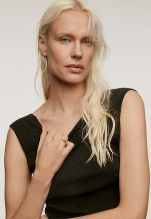 Mujer rubia con cabello largo, vistiendo una blusa sin mangas de punto oscuro, sosteniendo el escote con su mano izquierda y mostrando un anillo rojo en su dedo.