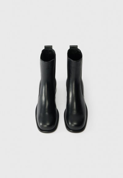 Melni ādas potītes zābaki ar gludu tekstūru, kvadrātveida purngalu un minimālu dizainu. Aprīkoti ar cilpām ērtai uzvilkšanai.