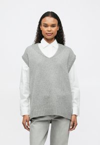 Γυναίκα φορώντας ένα λευκό πουκάμισο με γιακά κάτω από ένα γκρι πλεκτό αμάνικο πουλόβερ και ανοιχτό γκρι παντελόνι, στέκεται μπροστά σε λευκό φόντο.