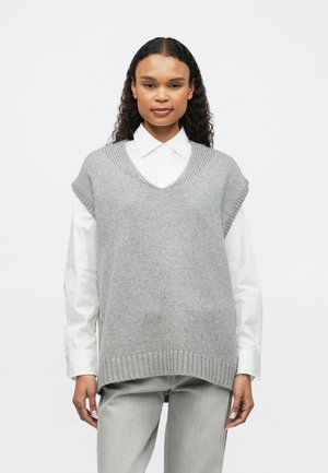 Femme portant une chemise blanche à col sous un pull sans manches gris tricoté et un pantalon gris clair, se tenant devant un fond blanc.