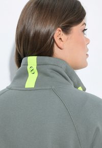 Sudadera gris con cuello alto, que presenta una franja de acento amarilla fluorescente y un logo circular en la parte posterior. Hecha de un suave tejido texturizado.