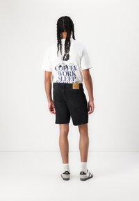 Un tricou alb cu mânecă scurtă, cu text bleumarin care spune "CAFEA MUNCĂ DORMI REPEATED", asortat cu pantaloni scurți negri din denim și sandale negre.