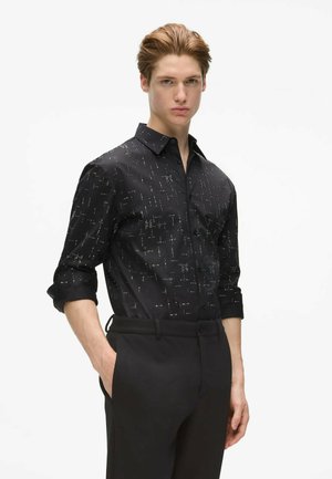 Jeune homme portant une chemise noire à motifs boutonnée avec les manches retroussées et un pantalon noir, posant avec une main dans la poche sur fond blanc.