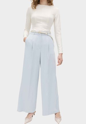 Frau trägt ein eng anliegendes, langärmeliges weißes Oberteil, das in hochtaillierte, weit geschnittene hellblaue Hosen mit einem dünnen beigen Gürtel und beigen Absätzen gesteckt ist.