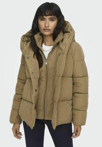 Hnědá péřová bunda s kapucí, s zipovým uzávěrem a velkými vypolstrovanými segmenty. Vyrobena z texturované tkaniny, nošena přes bílou košili.