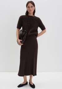 Donna che indossa un maglione corto a maniche corte marrone scuro e peloso, una gonna midi abbinata, ballerine nere, mentre tiene una pochette in pelle nera.