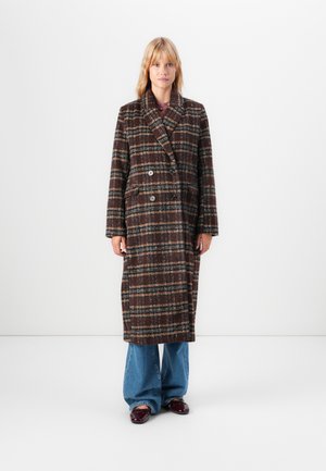Woman standing against white background wearing a long brown plaid double-breasted coat, wide-leg blue jeans, and burgundy shoes.