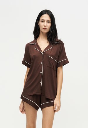 Femme aux longs cheveux foncés portant un haut brun à manches courtes boutonné et un short assorti avec des détails en passepoil blanc, debout devant un fond uni.