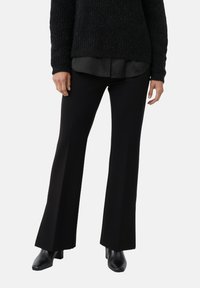 Kobieta ubrana w czarne rozszerzane spodnie, czarny teksturowany sweter na czarnej koszuli i czarne skórzane botki na obcasie na jednolitym tle.
