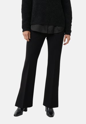 Kobieta ubrana w czarne rozszerzane spodnie, czarny teksturowany sweter na czarnej koszuli i czarne skórzane botki na obcasie na jednolitym tle.