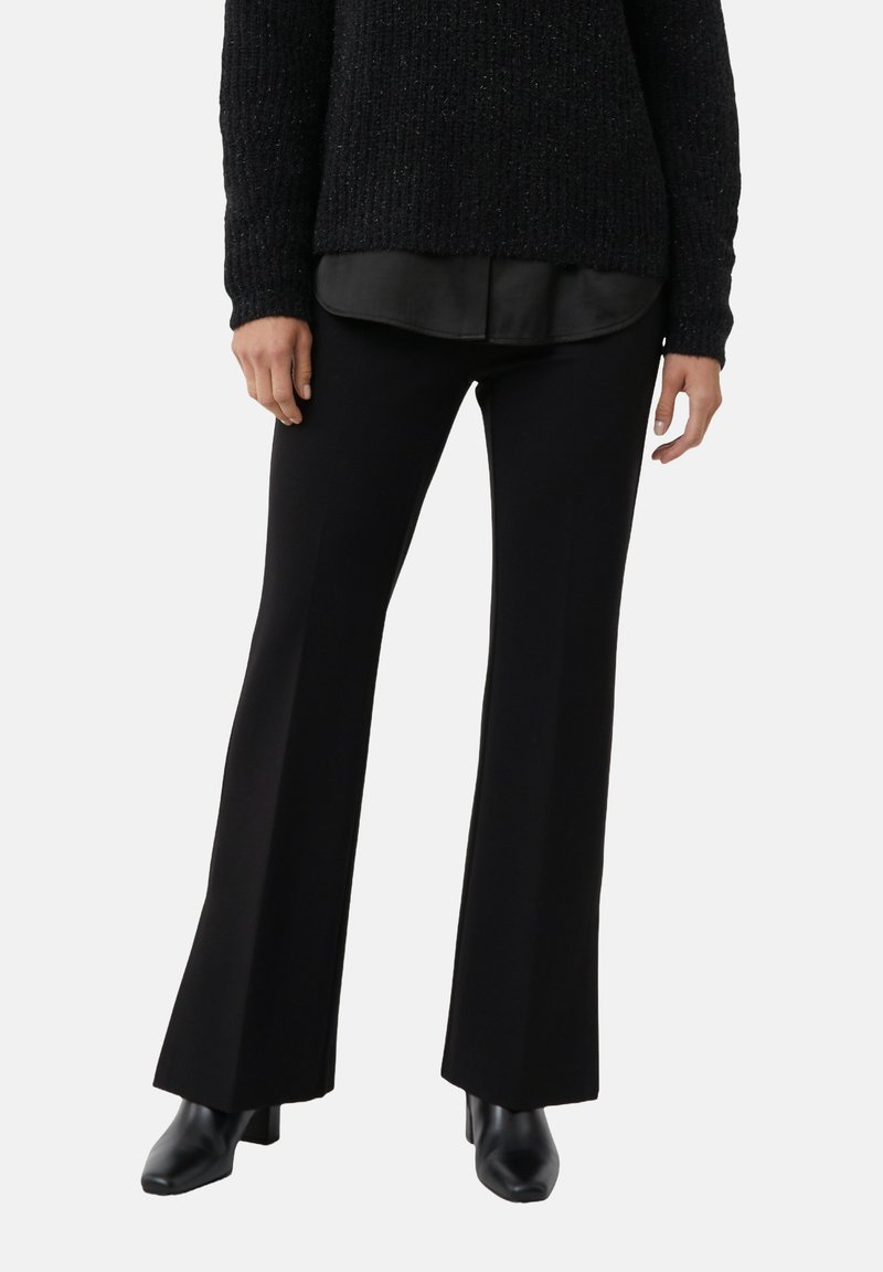 Kobieta ubrana w czarne rozszerzane spodnie, czarny teksturowany sweter na czarnej koszuli i czarne skórzane botki na obcasie na jednolitym tle.