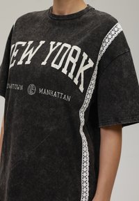 Svart oversized t-skjorte med "NEW YORK" grafikk i hvitt, med blondepaneldetalj på skuldrene. Mykt, teksturert stoff.