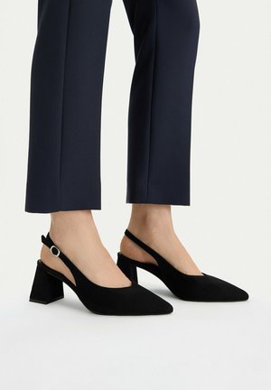 Donna con sandali neri con tacco largo e cinturino sul tallone, abbinati a pantaloni corti blu scuro, in piedi su una superficie bianca.