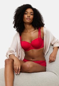 Conjunto de lingerie vermelha com um soutien levemente acolchoado e detalhes em renda, acompanhado de umas cuecas correspondentes. Usado por baixo de um robe aberto em tom bege suave.