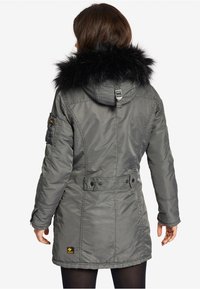 Vrouw in een grijze winterjas met een zwarte bont omzoomde capuchon, staande met de rug naar de camera tegen een witte achtergrond.