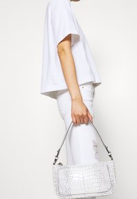 Bolso de mano embosado en piel de cocodrilo blanco con una correa corta, que presenta una textura suave y una forma rectangular, sostenido por una persona vestida de blanco.