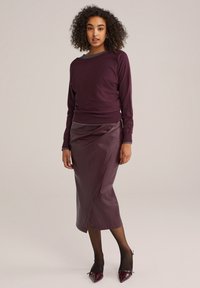 Burgunderfarbener Pullover mit langen Ärmeln und Rundhalsausschnitt, kombiniert mit einem bordeauxfarbenen Rock aus Kunstleder mit hoher Taille, der ein Wickeldesign und einen Schlitz aufweist.