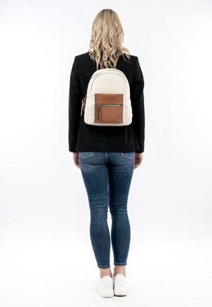 Femme portant une veste noire, un jean bleu, des baskets blanches et un sac à dos crème avec une poche avant marron, se tenant de dos sur fond blanc.