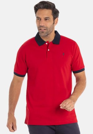 Homme avec de courts cheveux foncés portant une chemise polo rouge avec un col noir et des bords de manche noirs, se tenant devant un fond clair uni.