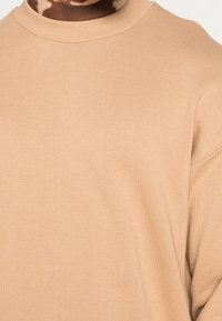 Béžový oversized svetr s ribovaným kulatým výstřihem, s hladkou texturou a volným střihem, zobrazený na světlém pozadí.