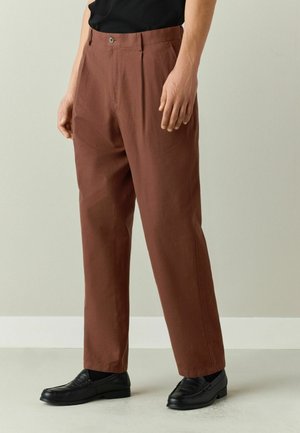 Man wearing brown straight-leg pants, black loafers, and black socks, standing against a plain light background.