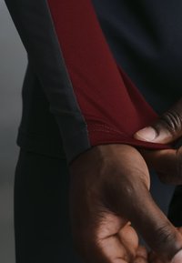 Κοντινό πλάνο ενός σκουρόχρωμου χεριού που ρυθμίζει την μανσέτα ενός ενδύματος με μακρύ μανίκι από κόκκινο και σκούρο γκρι ύφασμα.