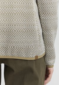 Pull en tricot texturé beige et crème avec un motif de diamant, doté d'un ourlet lisse et d'une étiquette en cuir en bas. Associé à un pantalon olive.