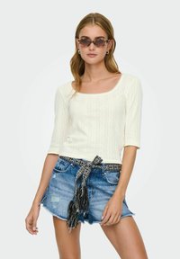 Witte geribde top met driekwartmouwen, gecombineerd met versleten denim shorts en een zwart-wit gemusterde riem met franjes.