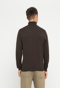 Bruine coltrui met lange mouwen, geribbelde kraag en manchetten, gemaakt van een zachte, geweven stof. G gedragen met lichtgekleurde broeken.