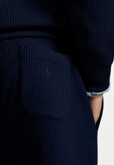 Tecido de malha em padrão waffle de cor azul-marinho com superfície texturizada, apresentando um bolso aplicado bordado com um logo e punhos enrolados numa cor contrastante.