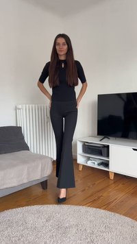 Kvinne med langt brunt hår står i svarte bukser og topp i et stue med sofa, radiator, TV og tregulv.