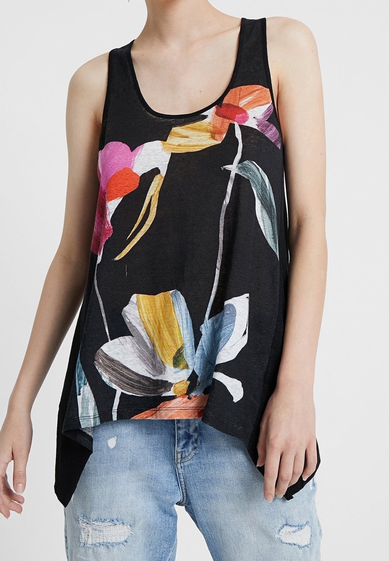 Femme portant un haut noir sans manches avec un grand imprimé floral coloré et un jean bleu clair déchiré, debout devant un fond uni.