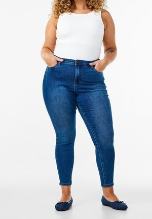 Krullendharige vrouw met een witte mouwloze top, hoog getailleerde blauwe skinny jeans en marineblauwe ballerina's, handen in de zakken.