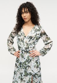 Φλοράλ φόρεμα με περιτύλιγμα, μακριά διάφανα μανίκια, ντεκολτέ σε σχήμα V και σκίσιμο μέχρι το μηρό. Διακοσμημένο με λευκό και πράσινο λουλουδάτο μοτίβο σε απαλό γαλάζιο φόντο.