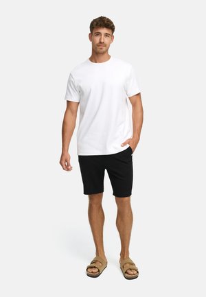 Mann steht frontal, trägt ein schlichtes weißes T-Shirt, schwarze Shorts und beige Sandalen vor hellgrauem Hintergrund.