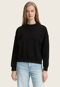 Felpa nera con scollatura ampia e spalle cadenti. Presenta maniche lunghe e una vestibilità comoda. Abbinata a jeans blu chiaro.