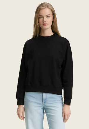 Sudadera negra con escote amplio y hombros caídos. Presenta mangas largas y un ajuste relajado. Combinada con denim de color azul claro.