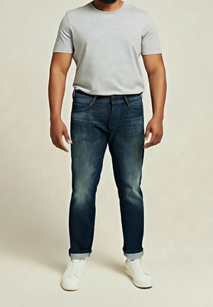 Mann trägt ein hellgraues T-Shirt mit kurzen Ärmeln, dunkelblaue hochgekrempelte Jeans und weiße Sneakers, steht vor einfarbigem Hintergrund.