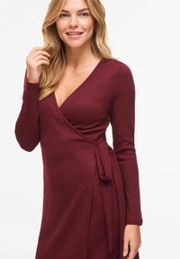 Robe portefeuille à manches longues en bordeaux, réalisée en tissu côtelé. Elle présente un col en V et un détail de nœud sur le côté à la taille.