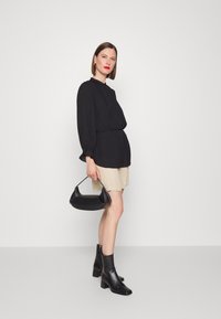 Blusa preta de mangas compridas com decote franzido, calções bege, botas de tornozelo pretas e uma pequena mala preta com um design curvado.