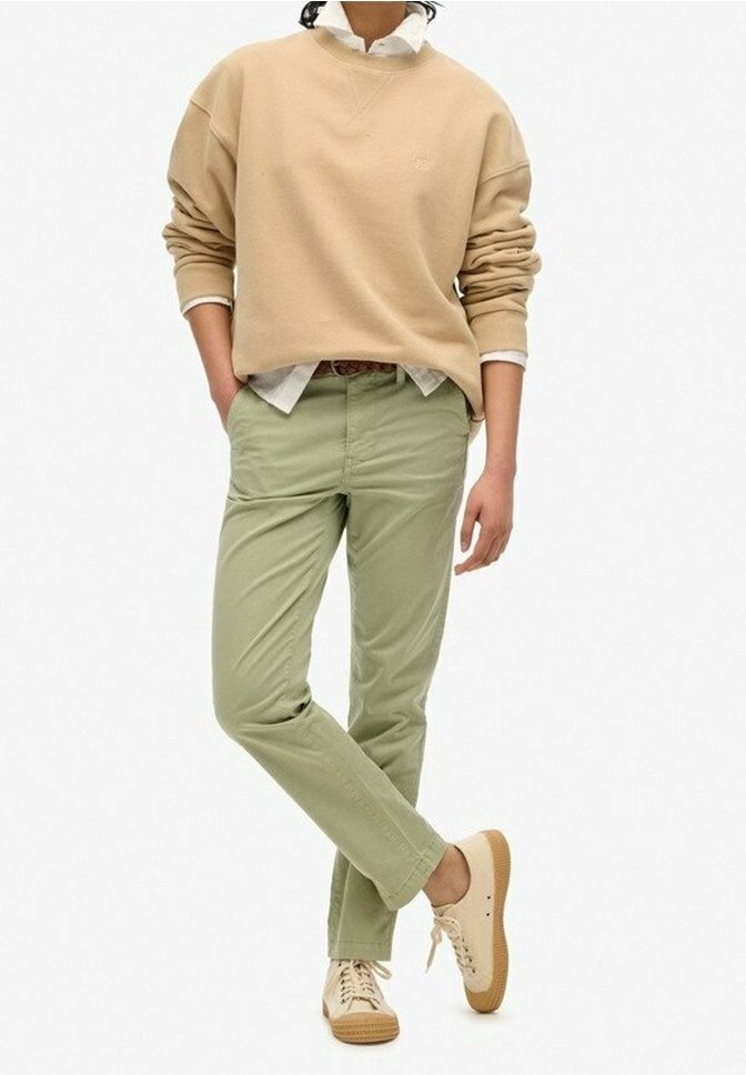 Personne portant un sweat-shirt beige, une chemise blanche, un pantalon vert clair et des baskets beiges, debout avec une jambe croisée sur l'autre.