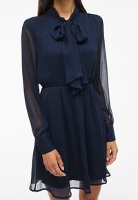 Γυναίκα που φοράει ένα διαφανές μπλε ναυτικό μακρυμάνικο φόρεμα με μια υφασμάτινη κομψή κορδέλα δεμένη στον γιακά και με ζωνάκι στη μέση.