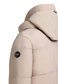 Beige polsterota jaka ar lielu kapuci un apaļu melnu logo uz pleca, ar gludu, nedaudz spīdīgu tekstūru.
