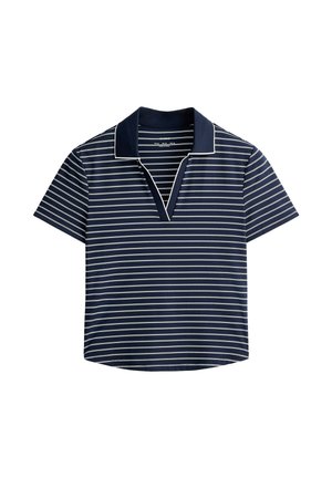 Femme portant un polo rayé bleu marine, une jupe plissée beige et une visière blanche, debout devant un fond gris uni.