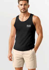 Schwarzes Tanktop mit dem Text "NEVER LESS®" in Weiß. Glatte Textur, runder Ausschnitt, kombiniert mit hellbeigen Shorts. Schlichtes Design.
