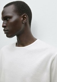 Hvid bomulds t-shirt med rund halsudskæring og let tekstur, bærer af en model med kort sort hår. Baggrunden er ensfarvet og lysefarvet.