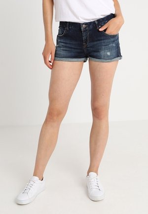 Person iført mørkeblå op rullede cowboyshorts, hvid t-shirt og hvide sneakers, der står med den ene hånd i lommen.