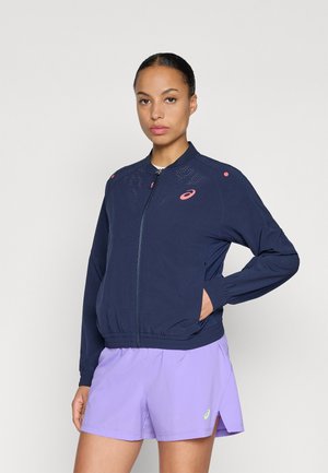 Marinejacke mit durchgehendem Reißverschluss und geripptem Saum, verziert mit rosa Akzentpunkten und durchbrochenen Designelementen. Tragen mit lilafarbenen Shorts.